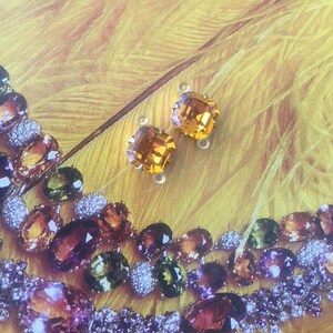 May include: Two square-cut, amber-colored gemstone earrings with silver-tone loops are displayed against a yellow background. A necklace with various gemstones in shades of amber, green, and purple is also visible.