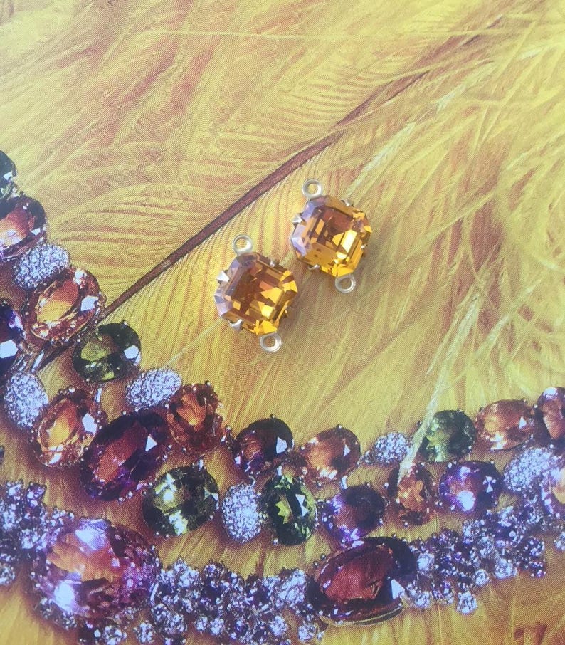 May include: Two square-cut, amber-colored gemstone earrings with silver-tone loops are displayed against a yellow background. A necklace with various gemstones in shades of amber, green, and purple is also visible.