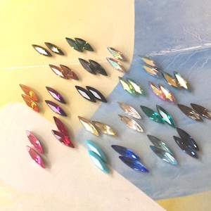 May include: An assortment of colourful, pointed, crystal-like rhinestones in various colours, including red, blue, green, and gold. The rhinestones are arranged in rows on a light blue and yellow surface.