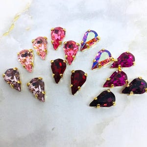Op de afbeelding: Een verzameling peervormige gefacetteerde kristallen in verschillende tinten roze, magenta en dieppaars, elk gezet in een goudkleurige metalen zetting. De kristallen vertonen verschillende niveaus van transparantie en iriserende afwerkingen.