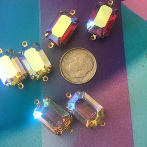 Könnte beinhalten: Sechs rechteckige, irisierende Kristall-Strasssteine mit goldfarbenen Metalleinfassungen. Die Kristalle haben einen Regenbogeneffekt, der Farben wie Rot, Gelb und Blau reflektiert. Eine US-Dime ist als Größenreferenz enthalten.