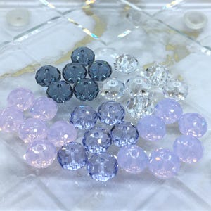 May include: A collection of faceted glass beads in various shades of blue, clear, and lavender. The beads are arranged on a clear, patterned surface, showcasing their reflective qualities. Ideal for jewelry making or craft projects.