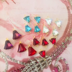 May include: A selection of colourful, triangular crystal pendants with gold loops. The pendants are in shades of blue, clear, red, and purple. The background is a soft pink with a decorative necklace.