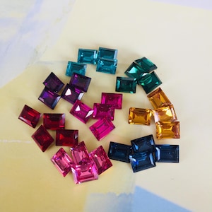 May include: Assortment of colorful rectangular gemstones in various shades of blue, green, purple, pink, and orange. The gemstones are arranged in a circular pattern on a light yellow surface.