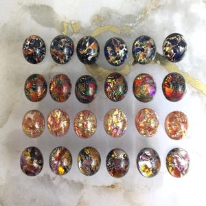 May include: A collection of twenty-four oval glass cabochons arranged in four rows of six. Each cabochon features a unique, multi-colored metallic foil inclusion pattern, displaying vibrant shades of blue, orange, gold, red, and purple.