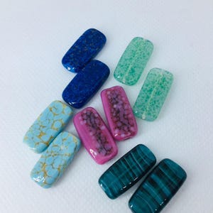 May include: A collection of rectangular glass beads in shades of blue, turquoise, pink, and green. Some beads feature textured patterns, while others have a smooth, striped design. The beads are displayed on a white background.