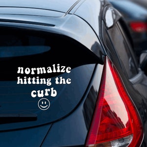 May include: A black car with a white vinyl sticker on the back window that says "normalize hitting the curb" with a smiley face below the text.