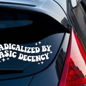 May include: White vinyl car decal with the text "RADICALIZED BY BASIC DECENCY" in a retro font, with star accents. The decal is affixed to the rear window of a black car.