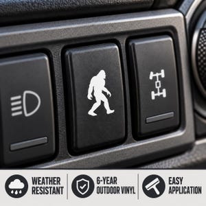 May include: A black rectangular vinyl decal featuring a white silhouette of a Bigfoot creature, designed to fit onto a vehicle dashboard button. The decal is weather resistant, made of 6-year outdoor vinyl, and is intended for easy application.