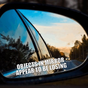 May include: A close-up of a car's side view mirror with a sticker that reads "Objects in mirror appear to be losing". The mirror reflects a blue sky with clouds and a road with trees on either side.
