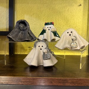 May include: A collection of ceramic ghost figurines. One is a dark gray, textured form. The others are white with various details: a green and red trimmed ghost, a ghost holding a pocket watch, and a ghost with a padlock.