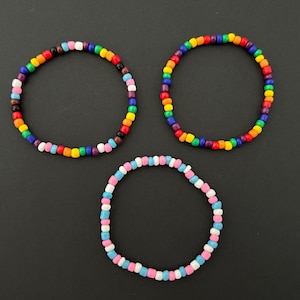 May include: Three beaded bracelets in a top-down view. Two bracelets feature a rainbow of colors, while the third bracelet has a pink, white, and blue color scheme. The bracelets are made of small, round beads.