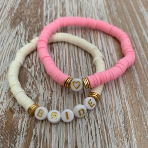 May include: Two bracelets: one in light pink and one in cream. The pink bracelet has a gold accent with a heart charm. The cream bracelet spells out the word "BRIDE" in gold letters on white beads. Both bracelets are on a wooden surface.
