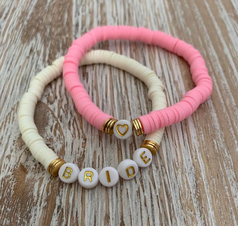 May include: Two bracelets: one in light pink and one in cream. The pink bracelet has a gold accent with a heart charm. The cream bracelet spells out the word "BRIDE" in gold letters on white beads. Both bracelets are on a wooden surface.