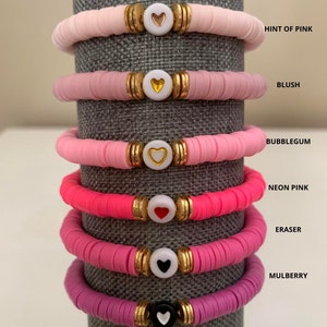 May include: Stack of six colorful bracelets. Each bracelet features a series of flat, round beads in shades of pink, with a heart-shaped charm in the center. The bracelets are labeled with the following colors: Hint of Pink, Blush, Bubblegum, Neon Pink, Eraser, and Mulberry.