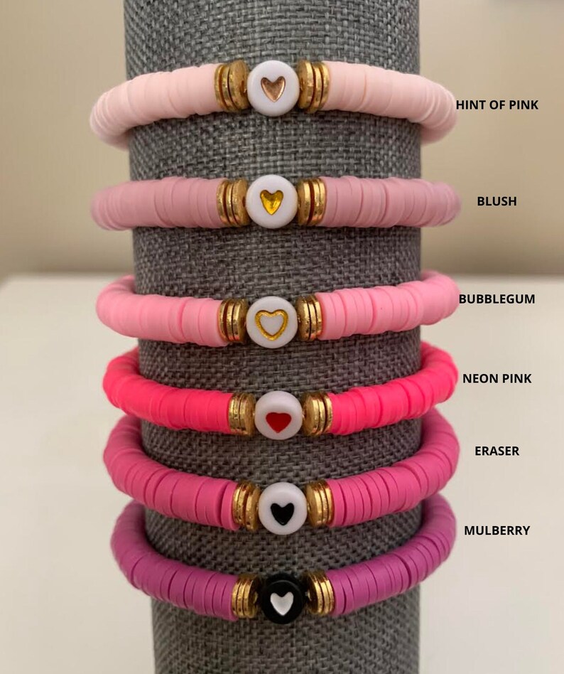 May include: Stack of six colorful bracelets. Each bracelet features a series of flat, round beads in shades of pink, with a heart-shaped charm in the center. The bracelets are labeled with the following colors: Hint of Pink, Blush, Bubblegum, Neon Pink, Eraser, and Mulberry.