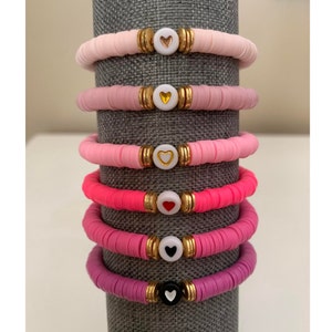 May include: Stack of six colorful bracelets. Each bracelet is made of flat, round beads in shades of pink and purple. Gold-colored spacers and a white heart-shaped bead are centered on each bracelet. The hearts are in various colors.