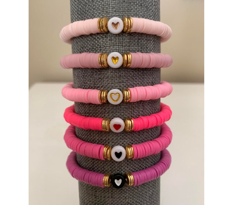 May include: Stack of six colorful bracelets. Each bracelet is made of flat, round beads in shades of pink and purple. Gold-colored spacers and a white heart-shaped bead are centered on each bracelet. The hearts are in various colors.