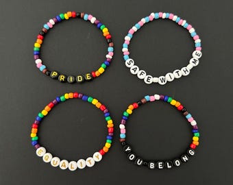 May include: Four beaded bracelets with rainbow-colored beads and letter beads spelling out "PRIDE", "SAFE WITH ME", "EQUALITY", and "YOU BELONG". The bracelets are on a dark gray background.