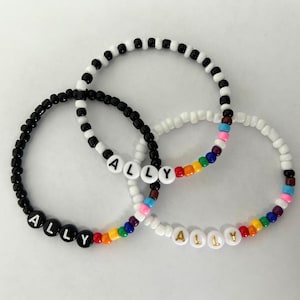 May include: Three beaded bracelets in black, white, and a mix of colors. Each bracelet spells "ALLY" with letter beads. One bracelet has a rainbow of beads. The bracelets are on a white background.