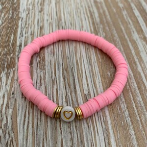 May include: A pink bracelet made of flat, round beads. The bracelet features a white bead with a gold heart design, accented by gold-colored spacers. The bracelet is displayed on a light wood surface.