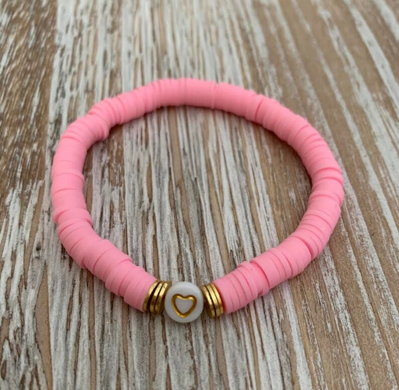 May include: A pink bracelet made of flat, round beads. The bracelet features a white bead with a gold heart design, accented by gold-colored spacers. The bracelet is displayed on a light wood surface.