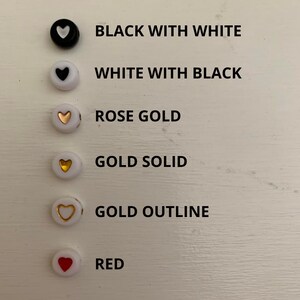 May include: A collection of round beads in various colors and designs. The beads include black with a white heart, white with a black heart, rose gold, gold solid, gold outline, and red. Each bead is displayed with its corresponding color description.