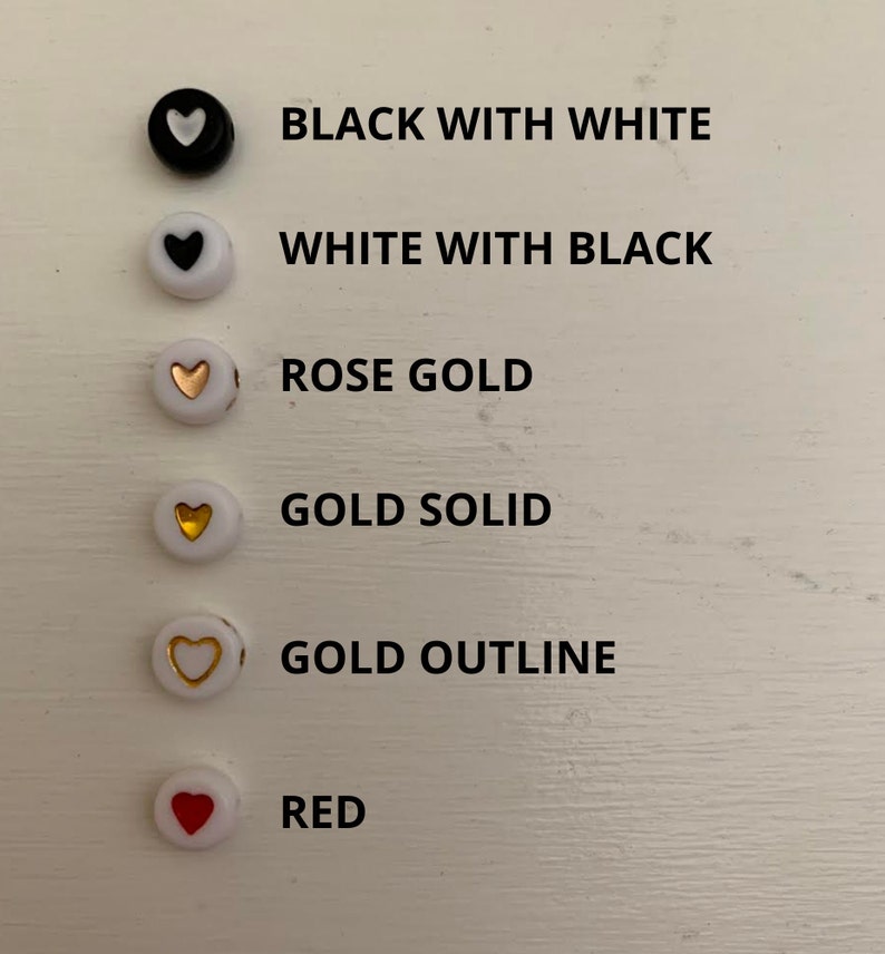 May include: A collection of round beads in various colors and designs. The beads include black with a white heart, white with a black heart, rose gold, gold solid, gold outline, and red. Each bead is displayed with its corresponding color description.