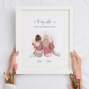 May include: A white framed print with a pink and white illustration of two women holding drinks. The text reads "To my sister" and "There's no better friend than a sister". The names "Karisa" and "Saraya" are written below the illustration.