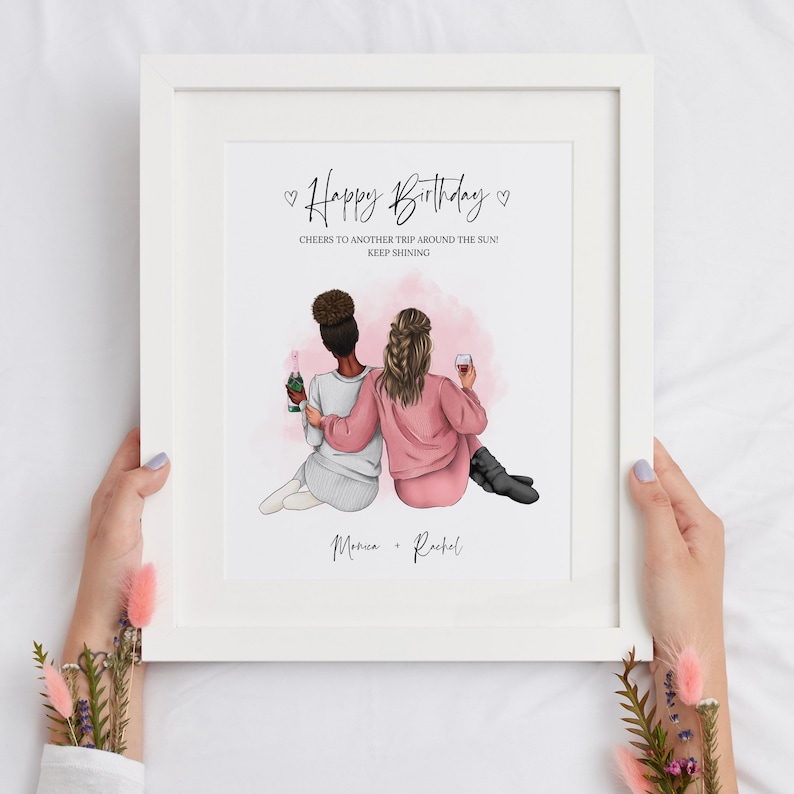 May include: A white framed print with a pink watercolor background featuring two women with brown hair holding glasses of wine. The text reads "Happy Birthday" and "Cheers to another trip around the sun! Keep shining." with the names "Monica + Rachel" at the bottom.