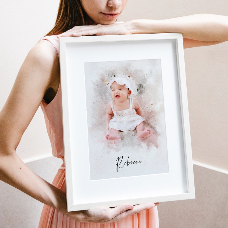 May include: A white framed print with a watercolor illustration of a baby girl wearing a white dress and a white flower crown. The baby is sitting on a pink and white background. The name "Rebecca" is written in black cursive at the bottom of the print.