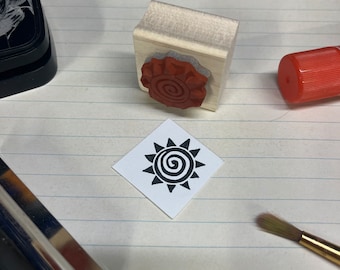 May include: A close-up of a wooden stamp with a red spiral design. The stamp is on a white lined paper surface with a black and white sun design stamp next to it. There is a pair of orange scissors, a roll of twine, a paintbrush, and a glue stick in the background.