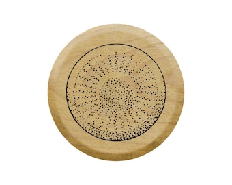 Op de afbeelding: Een ronde houten onderzetter met een zwart-witte grafische afbeelding van een zonnestralenontwerp.