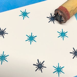 以下が含まれることがあります： 白いカードに、ターコイズブルーと黒の星形模様が複数印刷されています。画像の右上隅には、黒い星形模様のついた木製のスタンプが表示されています。