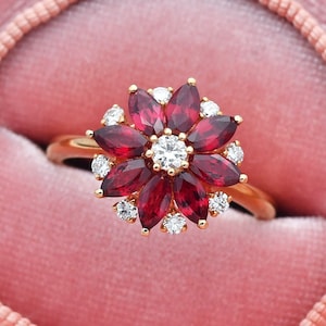 以下が含まれることがあります： ピンクのベルベットのリングボックスに置かれた、赤いルビーとダイヤモンドの花のデザインが施された金のリングです。