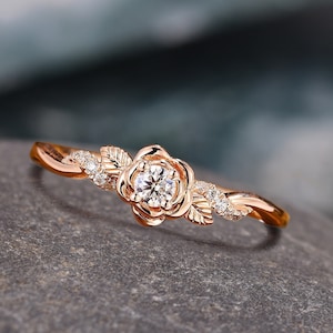 May include: Rose gold ring with a floral design. The ring features a central flower with a round, clear gemstone and smaller gemstones on the band. The band has a twisted design with leaf accents. The text "DreamsofDazzle" is at the top.