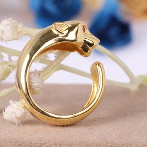 Puede incluir: Un anillo dorado con forma de pantera, con una gema roja como ojo. El anillo se curva, con la cabeza de la pantera en la parte superior. El anillo está sobre una superficie de color neutro con flores borrosas en el fondo.