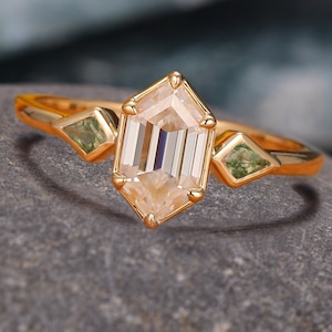 Puede incluir: Un anillo de oro con una piedra preciosa hexagonal de color marrón claro y dos piedras preciosas verdes más pequeñas en la banda.