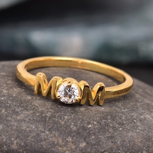 May include: Gold ring with the word "MOM" spelled out in raised letters with a small clear rhinestone in the center.