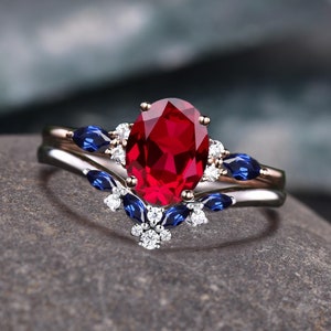 May include: A two-piece ring set featuring a large oval-cut ruby center stone. The engagement ring is accented with small white and blue gemstones. The band is silver and gold. The text "DreamsofDazzle" is at the top.
