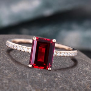 May include: A rose gold ring with a large, rectangular, deep red gemstone. The band is decorated with small, sparkling white gemstones. The ring is set on a grey stone surface.