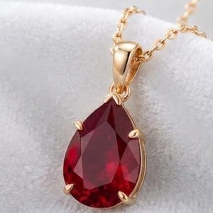 May include: A teardrop-shaped deep red gemstone pendant set in a gold-toned metal prong setting, suspended from a delicate gold-toned chain. The faceted stone reflects light, showcasing a rich crimson hue and a polished, reflective surface finish.