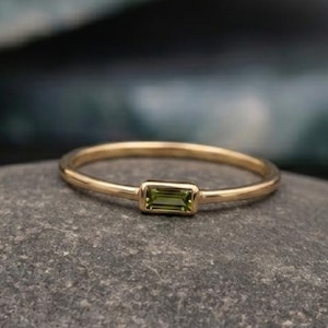 Puede incluir: Un delicado anillo de oro con una gema rectangular de color verde oliva. El anillo está colocado sobre una piedra gris, con un fondo verde y negro borroso. La banda del anillo es fina y pulida, resaltando el color de la gema.