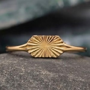 May include: A gold ring featuring a central rectangular plate with a sunburst pattern of engraved lines radiating from the center. The band is thin and smooth, connecting to the sides of the rectangular plate, creating a polished metallic finish.