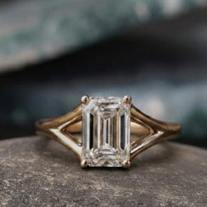 May include: A gold ring featuring a central emerald-cut clear gemstone. The band has a split-shank design that tapers towards the stone, creating a triangular opening on each side. The metal has a polished finish, and the stone is faceted.