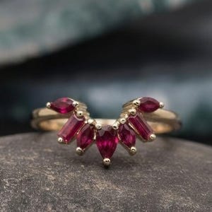 Op de afbeelding: Een gouden ring met een V-vormige chevronband bezet met vijf dieprode, peervormige robijnen. De stenen zijn in een symmetrisch waaiervormig patroon op de bovenkant van de gepolijste gouden band geplaatst, wat een opvallend puntig silhouet creëert.