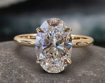 Puede incluir: Un anillo de compromiso de oro con un gran diamante de talla ovalada. El diamante es claro y multifacético, reflejando la luz. El anillo está sobre una piedra gris, con un fondo borroso.