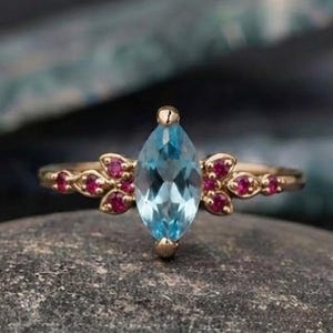 May include: A gold ring featuring a central marquise-cut light blue gemstone flanked by small, round, vibrant pink stones arranged in a floral pattern on each side of the band, creating a symmetrical design with a polished metal finish.