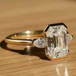 Puede incluir: Un anillo de oro con una gran piedra preciosa rectangular y transparente como pieza central. Dos piedras preciosas más pequeñas en forma de pera flanquean la piedra principal. El anillo está sobre una superficie de madera, con una sombra.