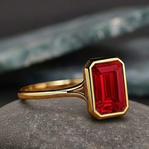 May include: A gold ring featuring a rectangular, bright red gemstone. The gemstone is set in a gold bezel, and the ring rests on a grey stone. The ring's band is a simple, polished gold.